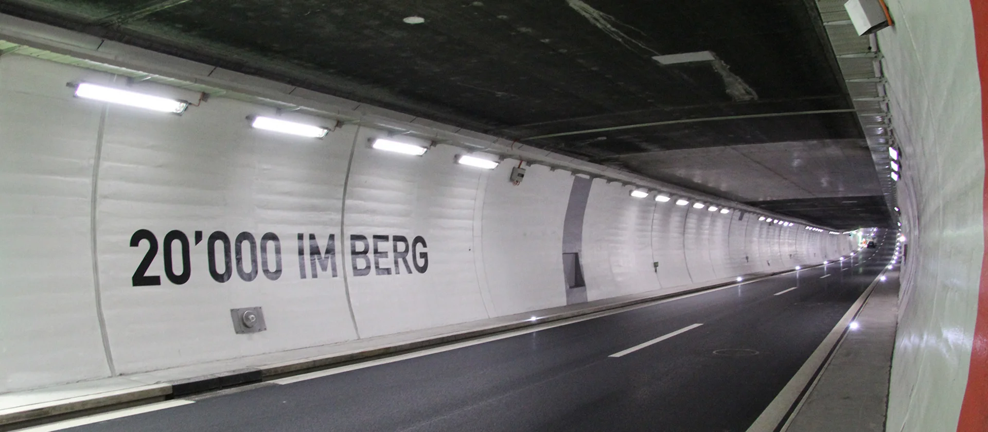 Foto: mit weissen Elementen verkleidete Wand im neuen Tunnel mit Aufschrift â20'000 im Bergâ und grauem Streifen; zweispurige, frisch asphaltierte Fahrbahn; Tunnelbeleuchtung