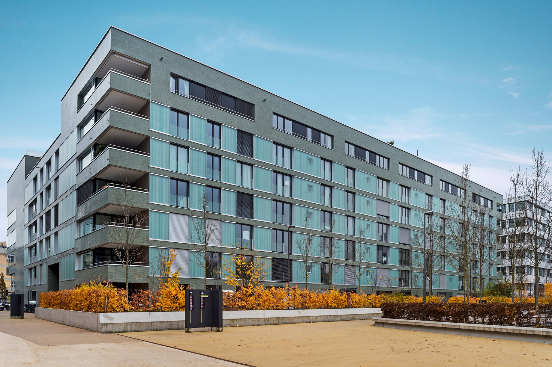 Photo : immeuble résidentiel moderne avec façades gris foncé et éléments turquoise ; devant, des aménagements extérieurs avec une végétation automnale luxuriante