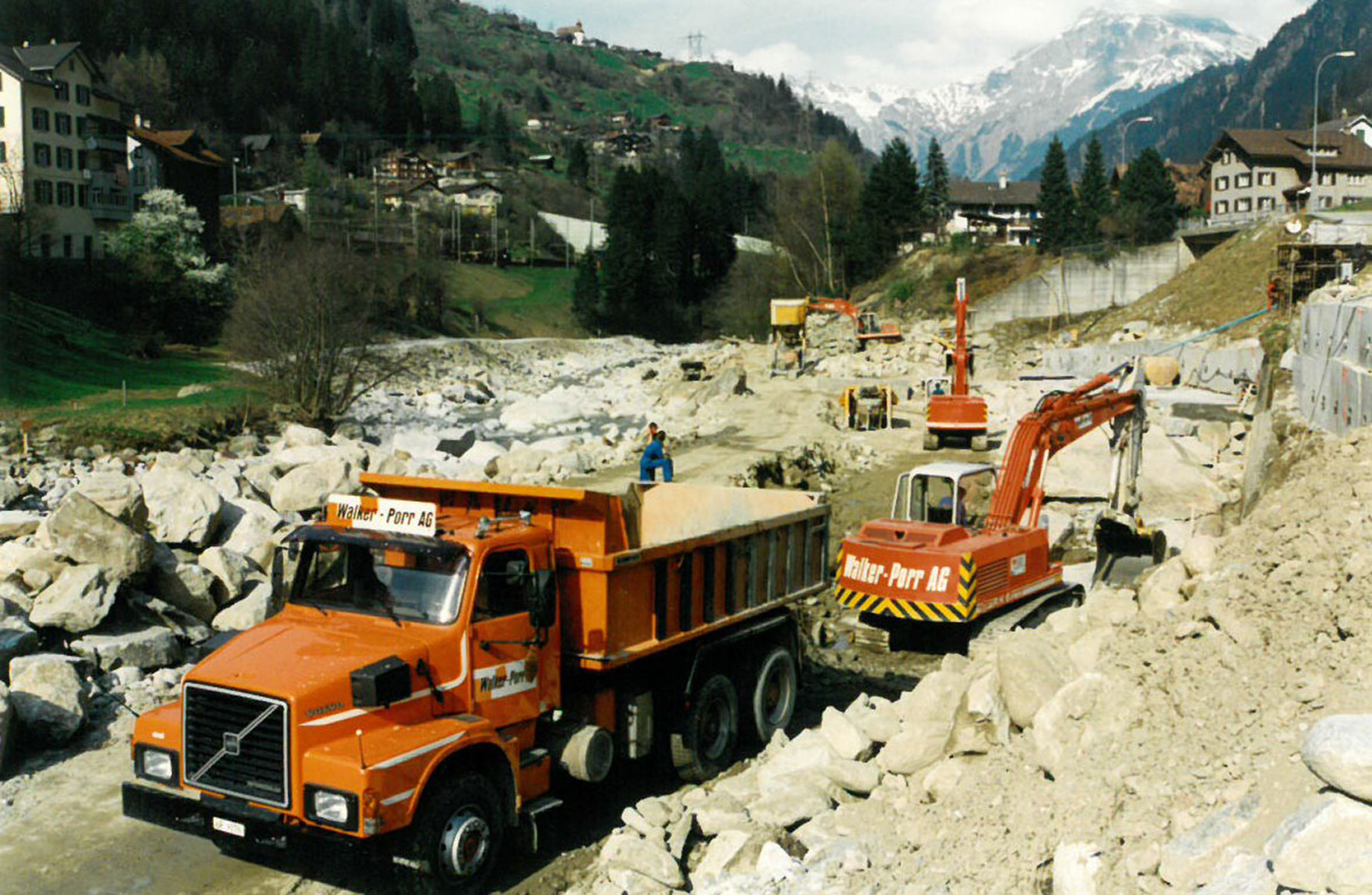 Photo: lancement historique d’un chantier de génie civil avec excavatrice et camions devant un paysage de montagne et de forêts.